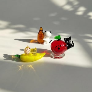 May include: A collection of miniature glass figurines. A yellow banana with a small figure, a brown bottle with a white cat figure, and a red apple with a black cat figure. The figurines are detailed and colorful.