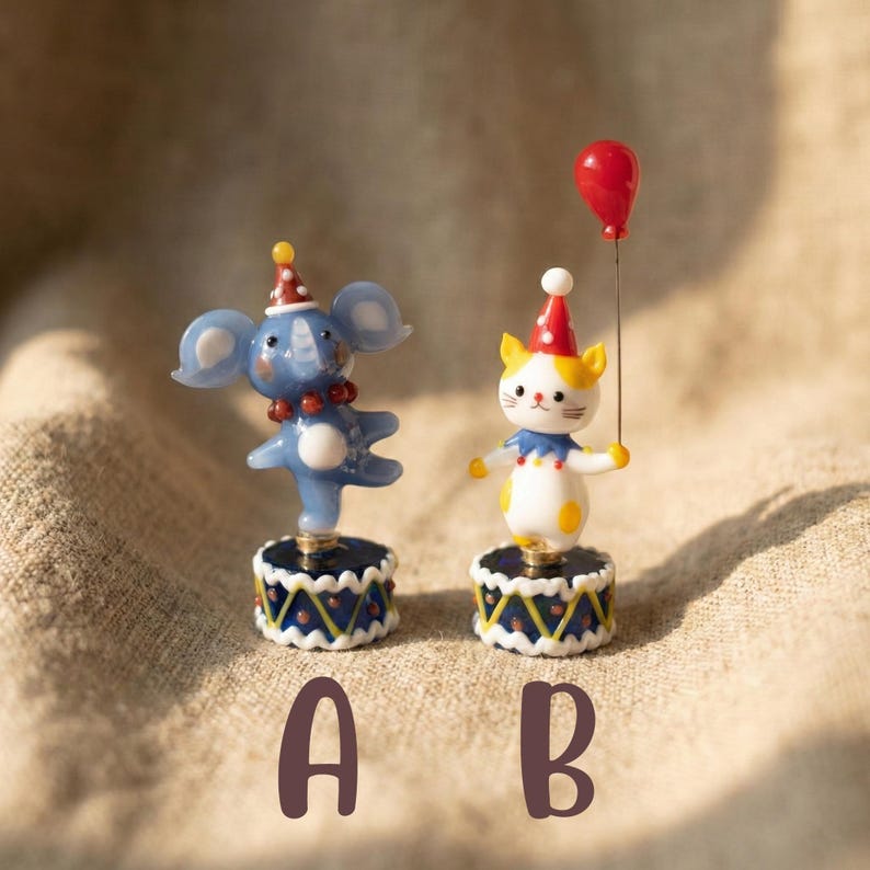 May include: Two whimsical figurines: a blue elephant and a white cat. The elephant wears a red party hat and a beaded necklace. The cat holds a red balloon. Both stand on decorative bases with blue and gold patterns. The letters "A" and "B" are below.