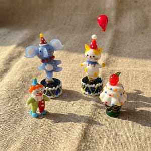 May include: A collection of miniature circus-themed figurines. Includes a blue elephant, a white and yellow cat, a clown, and a cupcake. Each figurine is on a decorative base and wears a party hat. A red balloon is also present.