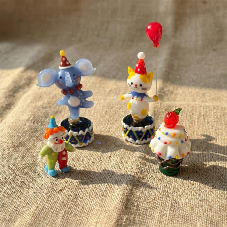 May include: A collection of miniature circus-themed figurines. Includes a blue elephant, a white and yellow cat, a clown, and a cupcake. Each figurine is on a decorative base and wears a party hat. A red balloon is also present.