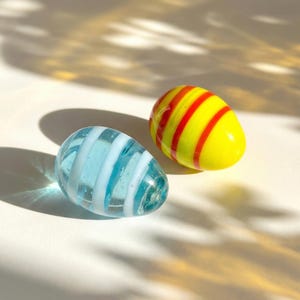Pode incluir: Dois ovos decorativos de vidro. Um é azul claro com listras brancas, o outro é amarelo com listras vermelhas. Os ovos são lisos e arredondados, com um acabamento brilhante. O fundo é branco com sombras.
