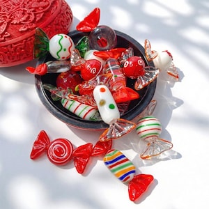 Peut inclure: Un assortiment de bonbons en verre colorés dans un bol noir et dispersés sur une surface blanche. Les bonbons sont rouges, blancs, verts et jaunes, avec divers motifs rayés et tourbillonnés. Une boîte rouge ornée est en arrière-plan.