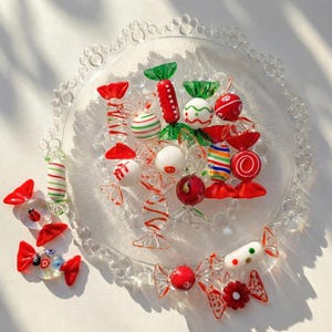 May include: A collection of colorful glass candies on a clear glass plate. The candies are various shapes and sizes, with red, green, white, and striped designs. The plate has a decorative beaded edge, and the candies are arranged on a white surface.