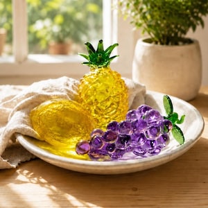 Pode incluir: Uma fruteira decorativa de vidro com um abacaxi amarelo, um limão amarelo e um cacho de uvas roxas. A fruta é disposta em um prato branco com um pano bege. O abacaxi tem um topo verde.