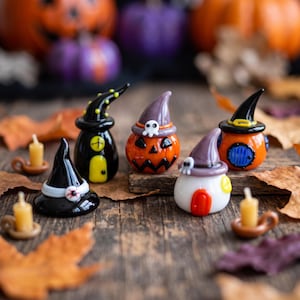 May include: A collection of miniature glass figurines featuring three small houses with pointed roofs, a black witch hat with an eyeball detail, and two tiny candles in brown holders. The houses include a black one, an orange pumpkin, and a white one.