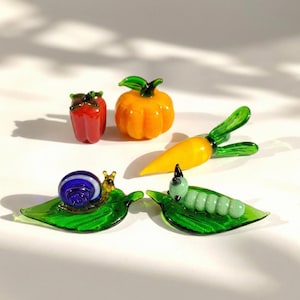 May include: A selection of glass ornaments featuring miniature vegetables and insects. The set includes a red pepper, an orange pumpkin, a yellow carrot, a snail with a blue shell, and a green caterpillar. The ornaments are on a white surface.
