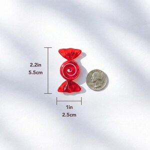May include: A red candy-shaped charm with a white swirl design, measuring 2.2 inches (5.5 cm) long and 1 inch (2.5 cm) wide. The charm is next to a U.S. quarter for size comparison. The candy has a wrapped appearance.