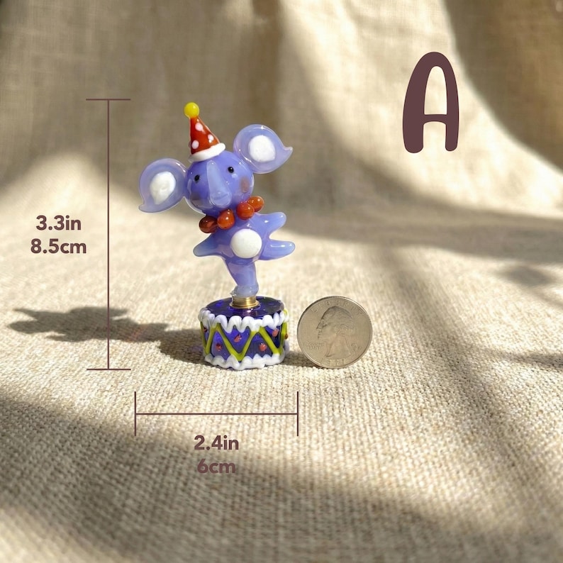 May include: A whimsical glass figurine of a purple elephant wearing a red and white party hat and a red beaded necklace. The elephant is balanced on a small, decorated platform. The figurine is 3.3 inches (8.5 cm) tall and the platform is 2.4 inches (6 cm) wide.