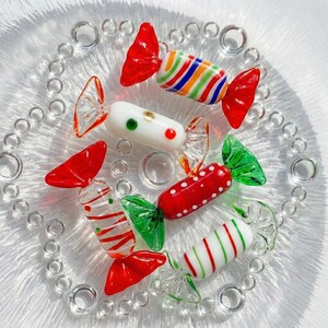 May include: A collection of colorful glass candies on a clear glass plate. The candies feature red, green, white, and multi-colored stripes and dots. The plate has a decorative bubble design around the edge.