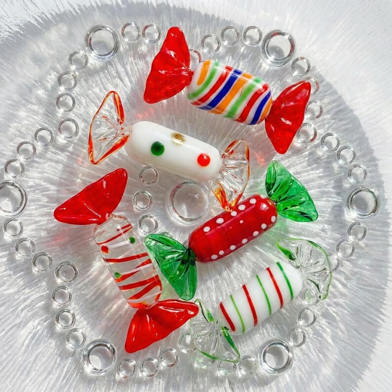 May include: A collection of colorful glass candies on a clear glass plate. The candies feature red, green, white, and multi-colored stripes and dots. The plate has a decorative bubble design around the edge.