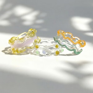 Può includere: Tre anelli di vetro colorati con bordi ondulati. Uno è giallo e rosa, un altro è trasparente con accenti gialli, verdi e blu, e il terzo è verde e arancione. Gli anelli sono esposti su una superficie bianca.