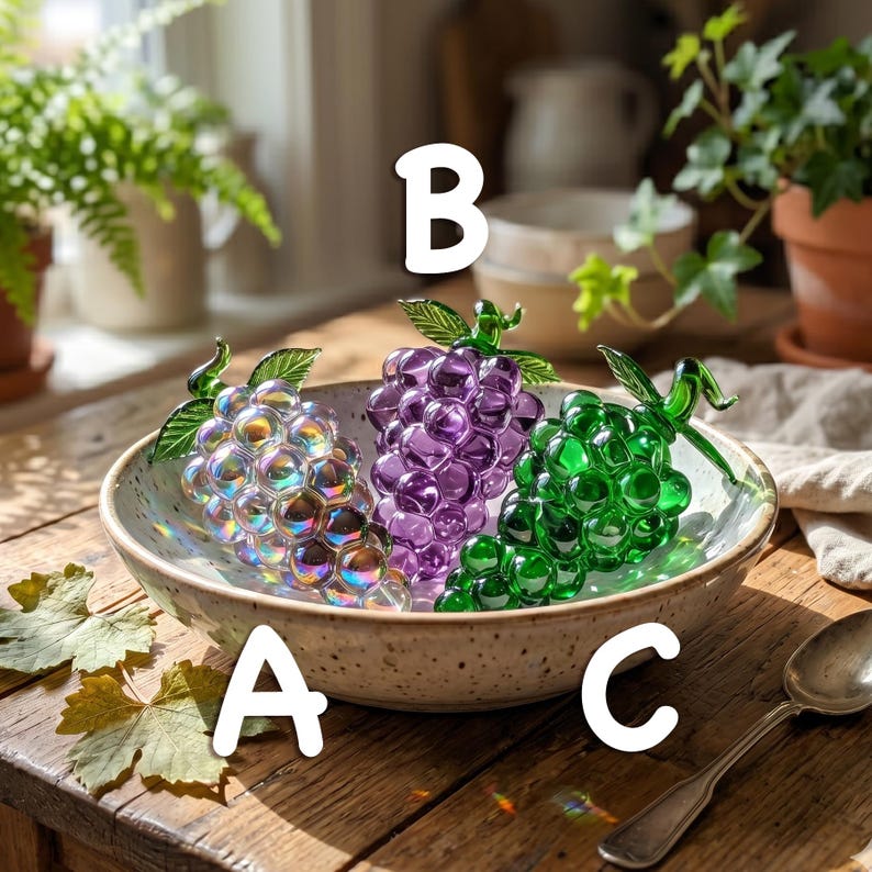 May include: Three glass grape clusters labeled A, B, and C inside a speckled ceramic bowl. Cluster A is iridescent, B is purple, and C is green. Each cluster features a green glass leaf and stem at the top, resting on a wooden surface.