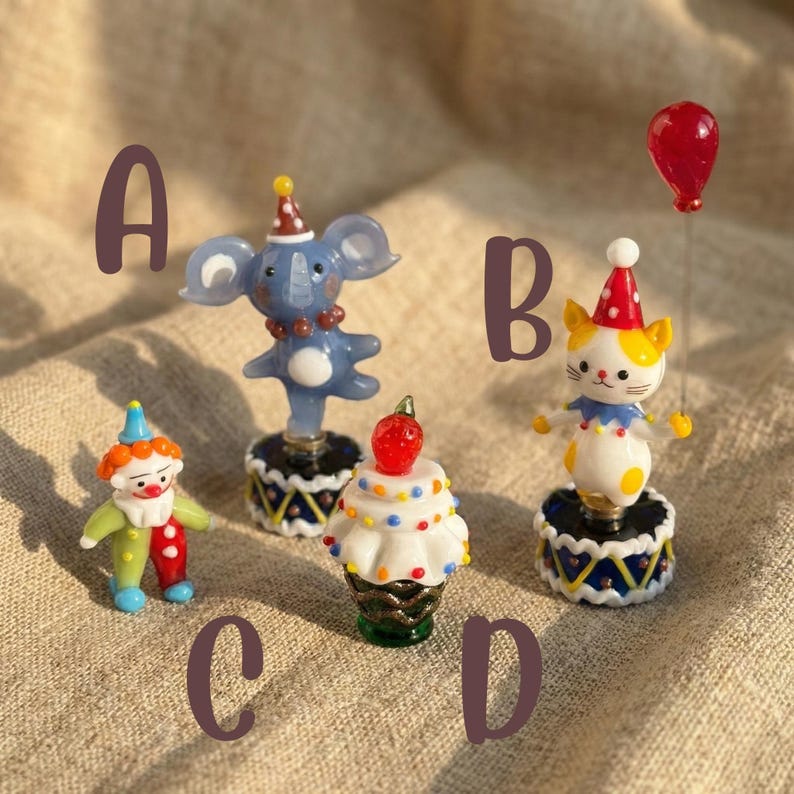 May include: A collection of miniature circus-themed figurines. The figurines include a blue elephant, a cat with a red party hat holding a red balloon, a clown, and a cupcake with a strawberry on top. Each figurine is on a small, decorative base.