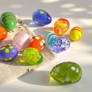 May include: A selection of decorative glass eggs in various colours and patterns. The eggs are in shades of green, blue, orange, yellow, and pink, with designs such as stripes and polka dots. They are displayed on a neutral-toned fabric.