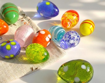 May include: A collection of colorful glass egg-shaped ornaments. The eggs feature various designs, including stripes, polka dots, and floral patterns, in shades of green, blue, orange, yellow, and pink. They are arranged on a neutral-colored fabric.