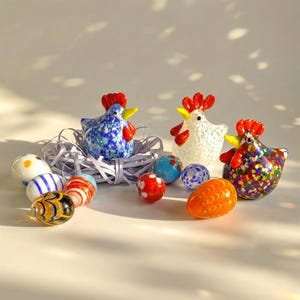 May include: Three glass hen figurines with red combs and yellow beaks, alongside decorative glass eggs. The hens are blue, white, and multicolored. The eggs feature various patterns and colors, including stripes, polka dots, and swirls. The figurines and eggs are arranged on a white surface.