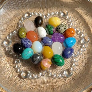 May include: A collection of polished gemstone eggs in various colors, including pink, blue, green, and yellow, arranged on a clear glass plate with a decorative beaded edge. The eggs are smooth and reflect light, showcasing their natural patterns.