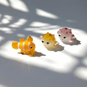 Peut inclure: Trois figurines de poissons en verre coloré. Un poisson-clown orange et blanc, et deux poissons-globes, un jaune et un rose. Tous ont des yeux noirs et de petites nageoires, présentés sur une surface blanche.