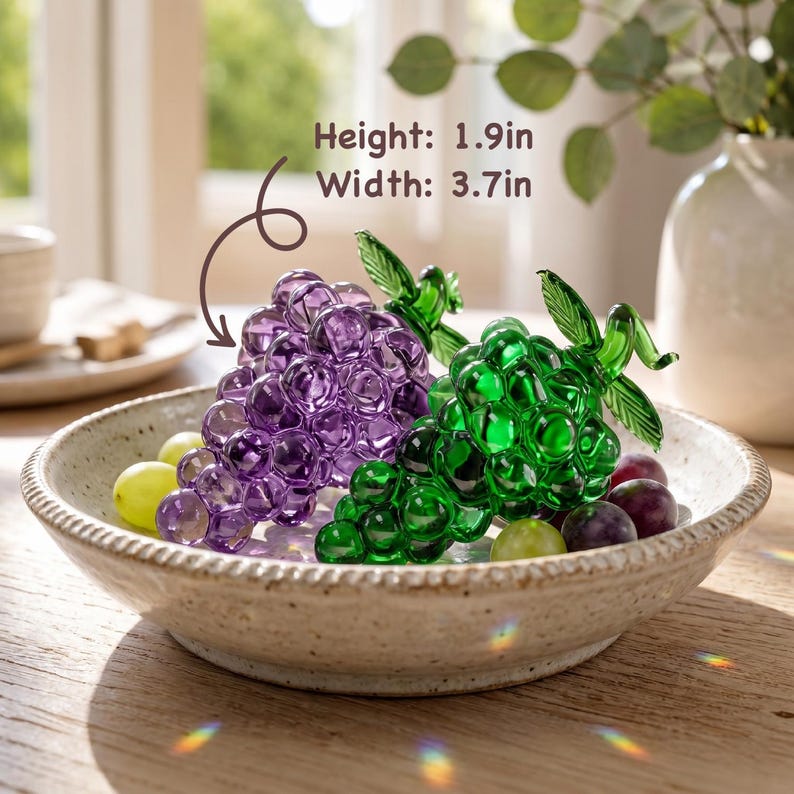 May include: Two glass grape clusters, one purple and one green, each with a green leaf. The text Height: 1.9in Width: 3.7in is visible. The grapes are in a speckled ceramic bowl with small loose grapes, resting on a wooden surface.