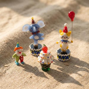 May include: A collection of miniature circus-themed figurines. Includes a clown, an elephant, a cupcake with a cherry, and a cat holding a red balloon. Each figurine is brightly colored and stands on a decorative base.