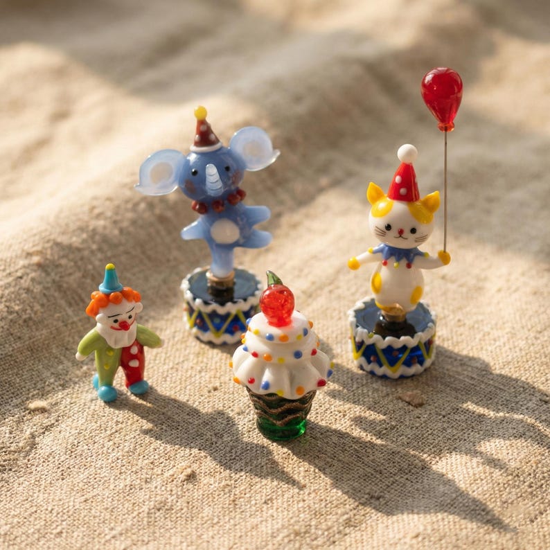 May include: A collection of miniature circus-themed figurines. Includes a clown, an elephant, a cupcake with a cherry, and a cat holding a red balloon. Each figurine is brightly colored and stands on a decorative base.