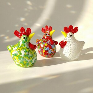 May include: Three decorative glass rooster figurines. One is green with yellow and white speckles, another is red with colorful speckles, and the third is white with a textured pattern. Each has a red comb and yellow beak.
