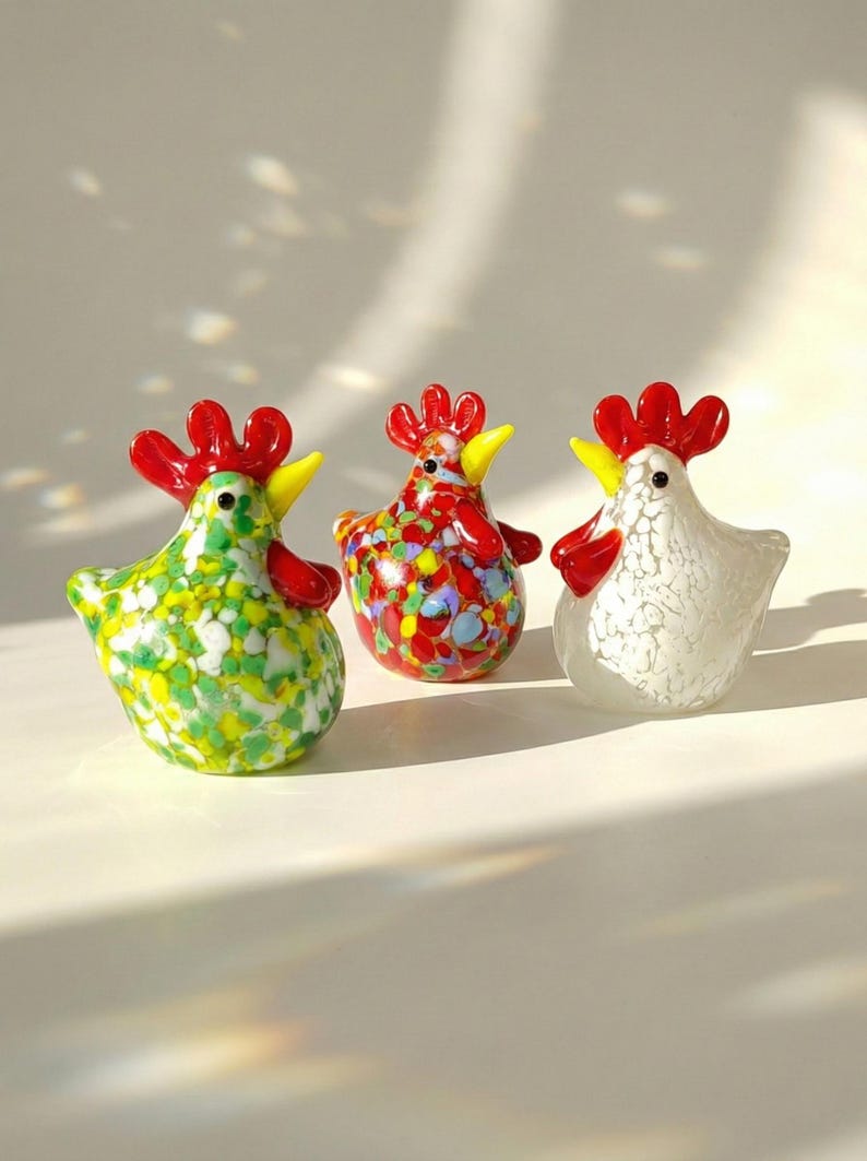 May include: Three decorative glass rooster figurines. One is green with yellow and white speckles, another is red with colorful speckles, and the third is white with a textured pattern. Each has a red comb and yellow beak.