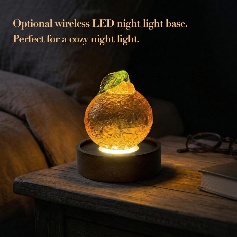 May include: A textured orange-shaped glass ornament with a green leaf on top, illuminated from below by a circular dark wooden base. The text "Optional wireless LED night light base. Perfect for a cozy night light." is visible in the upper left corner.