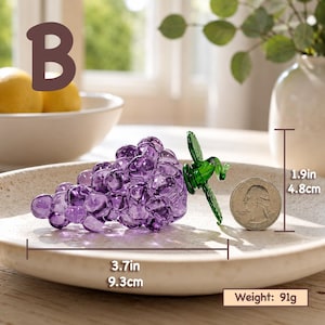 May include: A purple glass grape cluster figurine with a green glass leaf accent. The object measures 3.7 inches in length and 1.9 inches in height, with a weight of 91 grams. A US quarter coin is placed next to the glass grapes for scale.