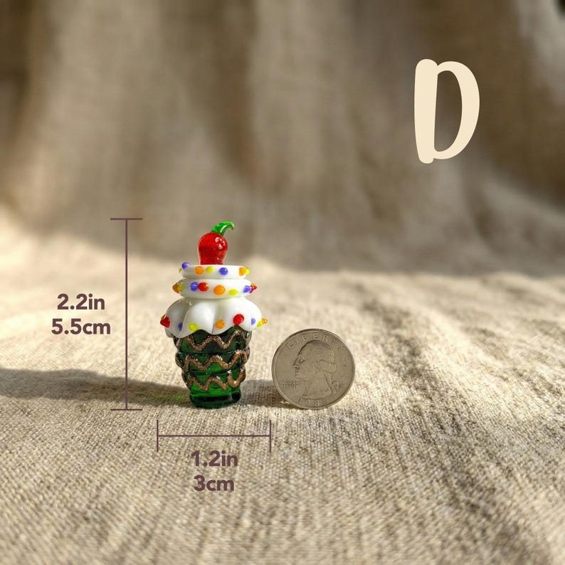 May include: A miniature glass cupcake figurine, 2.2 inches (5.5 cm) tall and 1.2 inches (3 cm) wide, with a green base, white frosting, and a red cherry on top. The frosting is decorated with colorful dots. A quarter is next to the cupcake.