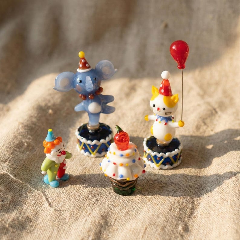 May include: A collection of miniature circus-themed figurines. Includes a blue koala, a clown, a cat holding a red balloon, and a cupcake with a cherry on top. Each figurine is on a decorative base with a blue and white pattern.