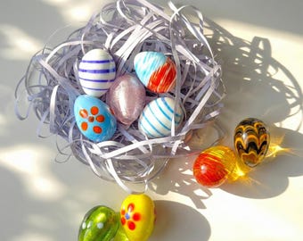 May include: A collection of decorative glass Easter eggs in various colors and patterns. Some eggs are nestled in a nest of shredded paper. The eggs feature designs such as stripes, swirls, and floral motifs, adding a festive touch.