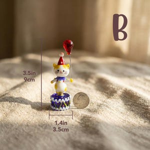 May include: A whimsical figurine of a cat wearing a party hat and holding a red balloon. The cat is 3.5 inches tall and stands on a 1.4-inch base. The figurine has yellow, white, blue, and red accents. A quarter is next to the figurine.