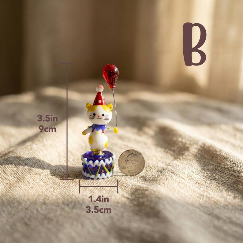 May include: A whimsical figurine of a cat wearing a party hat and holding a red balloon. The cat is 3.5 inches tall and stands on a 1.4-inch base. The figurine has yellow, white, blue, and red accents. A quarter is next to the figurine.
