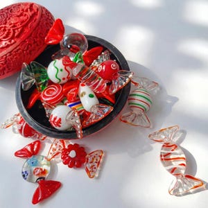 May include: A collection of colorful glass candies in a black bowl, with a red carved container in the background. The candies are red, white, and green, with various designs. Some candies are outside the bowl, scattered on a white surface.