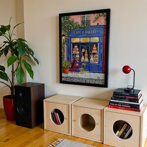 Charming Paris café art print featuring a classic blue storefront on a quaint city street, with a small dog waiting patiently beside a table of pastries. Croissants and coffee add a cozy morning feel, while soft light and European details evoke a relaxed Parisian atmosphere. Elegant yet whimsical wall art perfect for kitchens, cafés, or living spaces, celebrating travel, food, and everyday moments in France.