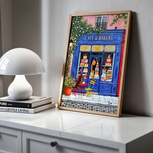 Charming Paris café art print featuring a classic blue storefront on a quaint city street, with a small dog waiting patiently beside a table of pastries. Croissants and coffee add a cozy morning feel, while soft light and European details evoke a relaxed Parisian atmosphere. Elegant yet whimsical wall art perfect for kitchens, cafés, or living spaces, celebrating travel, food, and everyday moments in France.