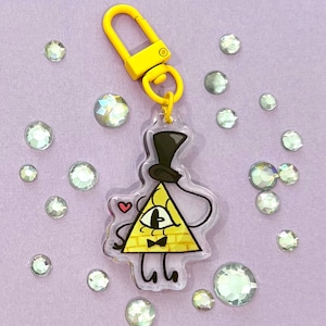 May include: A yellow and black cartoon character keychain with a yellow clasp. The character is a yellow triangle with a top hat, bow tie, and one eye. The keychain is surrounded by clear, sparkling rhinestones on a purple background.