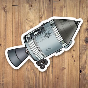 May include: A sticker featuring a detailed illustration of a spacecraft, predominantly in shades of gray and black. The design includes intricate details of the spacecraft's components, with a white border. The sticker is placed on a wooden surface.