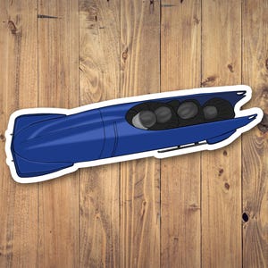 May include: A sticker featuring a blue bobsled with a white border. The bobsled has a streamlined design with four black helmeted figures visible inside. The sticker is on a wooden background.