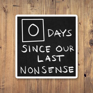 May include: A black chalkboard with white text that reads "0 DAYS SINCE OUR LAST NONSENSE." The number 0 is inside a white square. The chalkboard has a white border and is set against a wooden background.