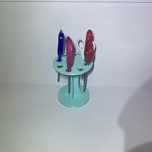 Könnte beinhalten: Ein hellblauer Scherenhalter mit mehreren Scherenpaaren in verschiedenen Farben. Die Scheren sind blau, rosa, weiß und rot. Der Halter hat eine kreisförmige Oberseite mit Löchern für die Scheren und einen runden Sockel.