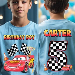 May include: Light blue t-shirt with the text "BIRTHDAY BOY" on the front and "CARTER" and the number "5" on the back. The front features a red race car graphic, checkered flags, and a trophy. The back also has a race car graphic.