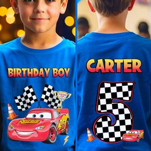 May include: Blue t-shirt with a graphic of Lightning McQueen from the movie Cars. The front of the shirt says "BIRTHDAY BOY" and features race flags, a trophy, and a lightning bolt. The back of the shirt says "CARTER" and "5" with a checkered pattern.