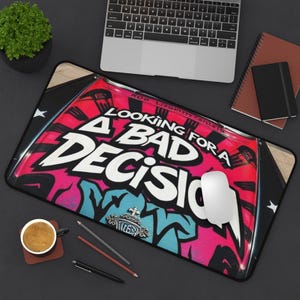 以下が含まれることがあります： ピンクと青のグラフィティデザインの上に、白い文字で「LOOKING FOR A BAD DECISION」と書かれた長方形のマウスパッド。白いコンピューターマウスがパッドの上にあり、背景にはラップトップとコーヒーカップがあります。