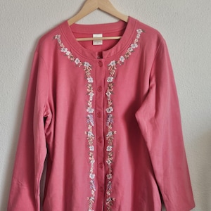 May include: A coral-colored button-up cardigan with floral embroidery. The embroidery features white, purple, and green flowers along the neckline and down the front. The long-sleeved cardigan is hanging on a wooden hanger.