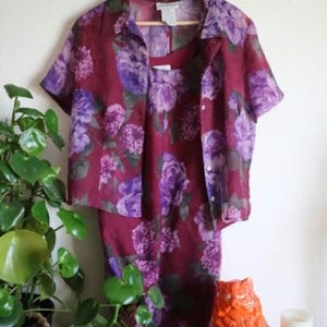 May include: A burgundy floral print outfit, consisting of a short-sleeved button-up shirt and a matching tank top and skirt. The floral pattern features purple and lavender flowers. The outfit is hanging on a wooden hanger.