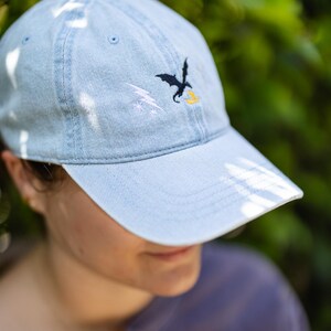 May include: A light blue denim baseball cap featuring an embroidered design on the front panel. The embroidery consists of a black dragon silhouette, a white lightning bolt, and a small yellow flame shape positioned below the dragon.