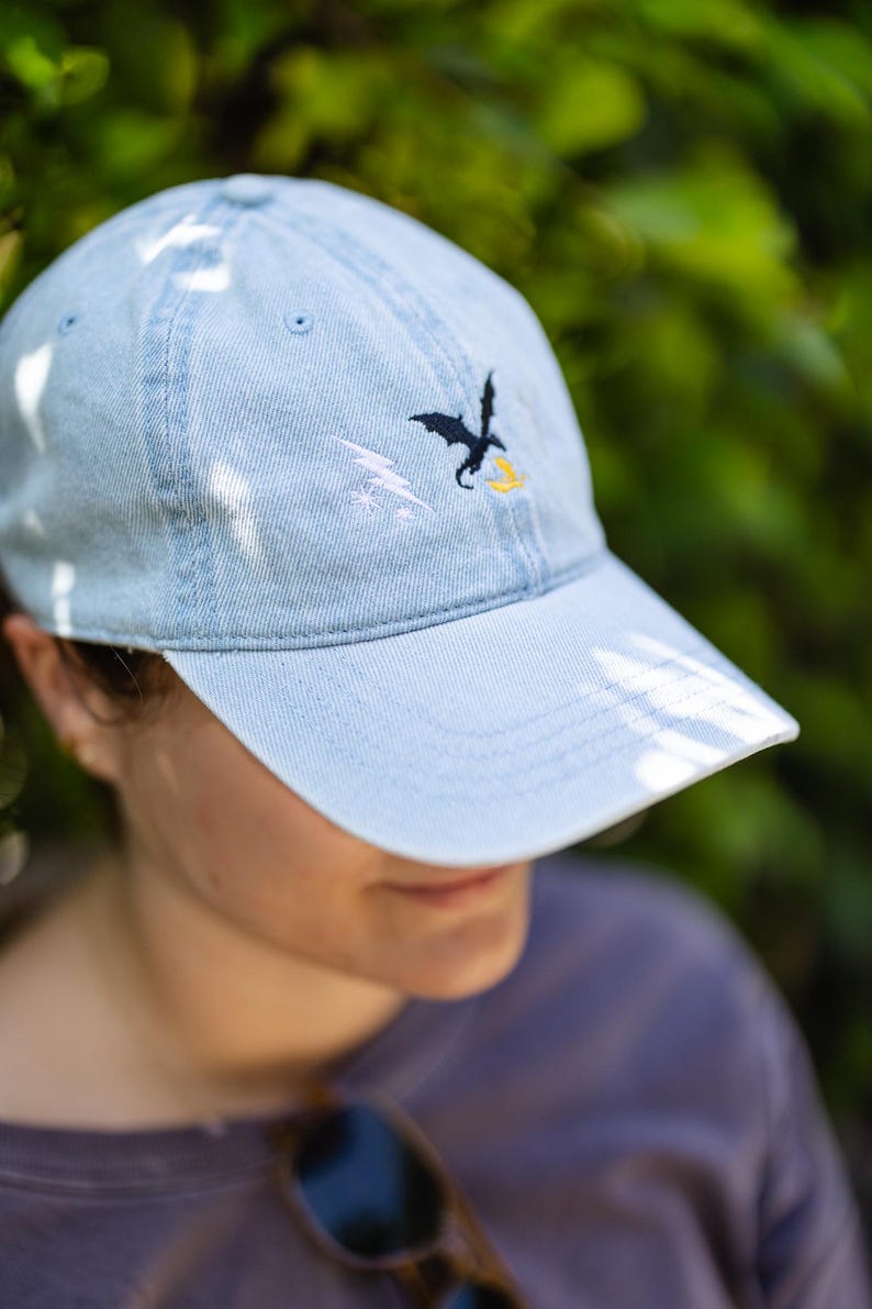 May include: A light blue denim baseball cap featuring an embroidered design on the front panel. The embroidery consists of a black dragon silhouette, a white lightning bolt, and a small yellow flame shape positioned below the dragon.