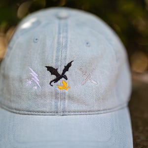 May include: A light blue denim baseball cap featuring three embroidered designs on the front panel: a white lightning bolt with a star, a black dragon silhouette above a small yellow flame, and two crossed silver swords.